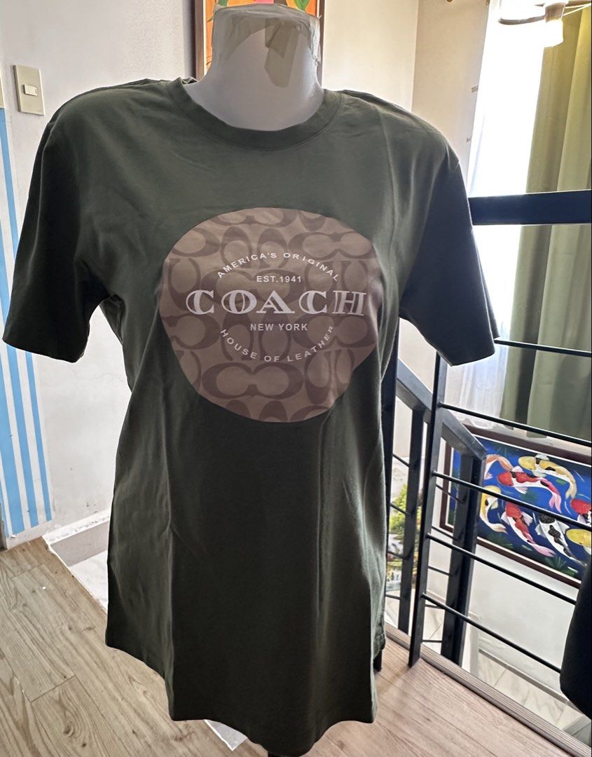 Coach Green Graphic Tee (Category: Shirts) located in Manila. Submitted by rachael_23 (Listing ID 1469083403, Image 1). Description: - Brand: Coach - Color: Dark Green - Material: 100% Cotton - Fit: Regular fit with round neckline and short sleeves - Design: Large circular graphic print with Coach New York text and signature monogram pattern - Back: Solid dark green - Condition: Clean fabric, no stains, holes, or damage found - Style: Great basic