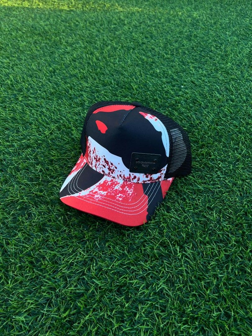 Dolce & Gabbana Red Black Splatter Trucker Cap (Category: Caps & Hats) located in Pasig City. Submitted by paulclzdo (Listing ID 1469063987, Image 2). Description: - Brand: Dolce & Gabbana - Style: Trucker cap with curved brim and snapback closure - Design: Eye-catching red, black, and white splatter pattern on front panels - Details: Black branded leather patch on the front right, white embroidered D&G lettering above the back strap - Comfort & 
