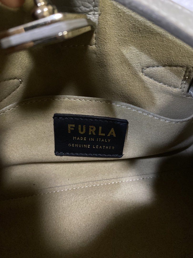 Furla bag (Category: Purses & Pouches) located in San Antonio. Submitted by clinicallycute_. (Listing ID 1469047571, Image 3). Description: - Brand: Furla - Material: Genuine leather in neutral beige - Hardware: Elegant gold accents with front strap closure - Design: Structured silhouette with a single top handle - Interior: Fabric-lined with 'Made in Italy' and 'Genuine Leather' brand patch - Size: Compact, ideal for daily essentials - Condition