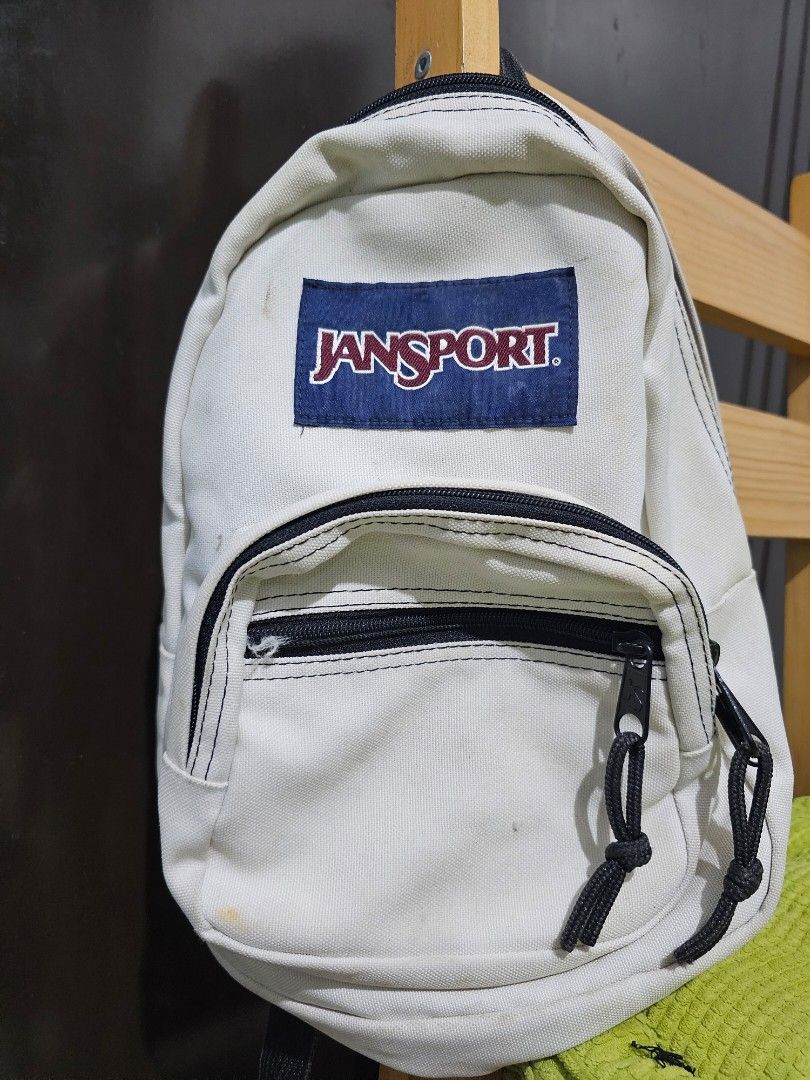 JanSport Mini White Backpack (Category: Backpacks) located in Dasmarinas. Submitted by jazvielle (Listing ID 1469087614, Image 2). Description: - Authentic JanSport white mini backpack - Color: White with black zippers, rope pulls, and adjustable shoulder straps - Features: Front zipped pocket and classic blue and red woven logo patch - Size: Compact, roughly 12 inches tall - Condition: Pre-loved with visible dirt marks and scuffs on the canvas f
