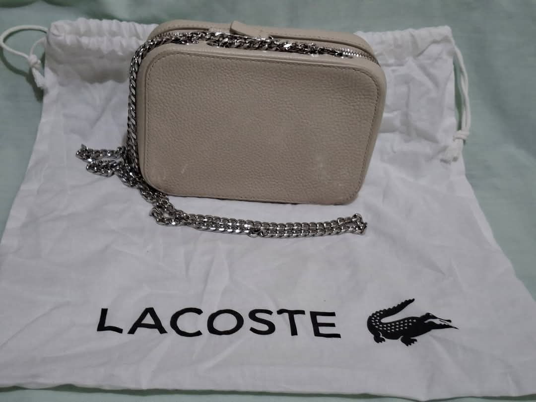 Lacoste Cream Camera Bag with Silver Chain (Category: Cross-body Bags) located in Manila. Submitted by closetprincess (Listing ID 1468957986, Image 3). Description: - Brand: Lacoste - Material: Cream pebbled leather - Design: Large embossed crocodile logo on front with silver-tone chain strap and hardware - Closure: Top zip closure with fabric interior lining - Inclusions: Original white dust bag and paper tag - Condition: Excellent, well-kept.