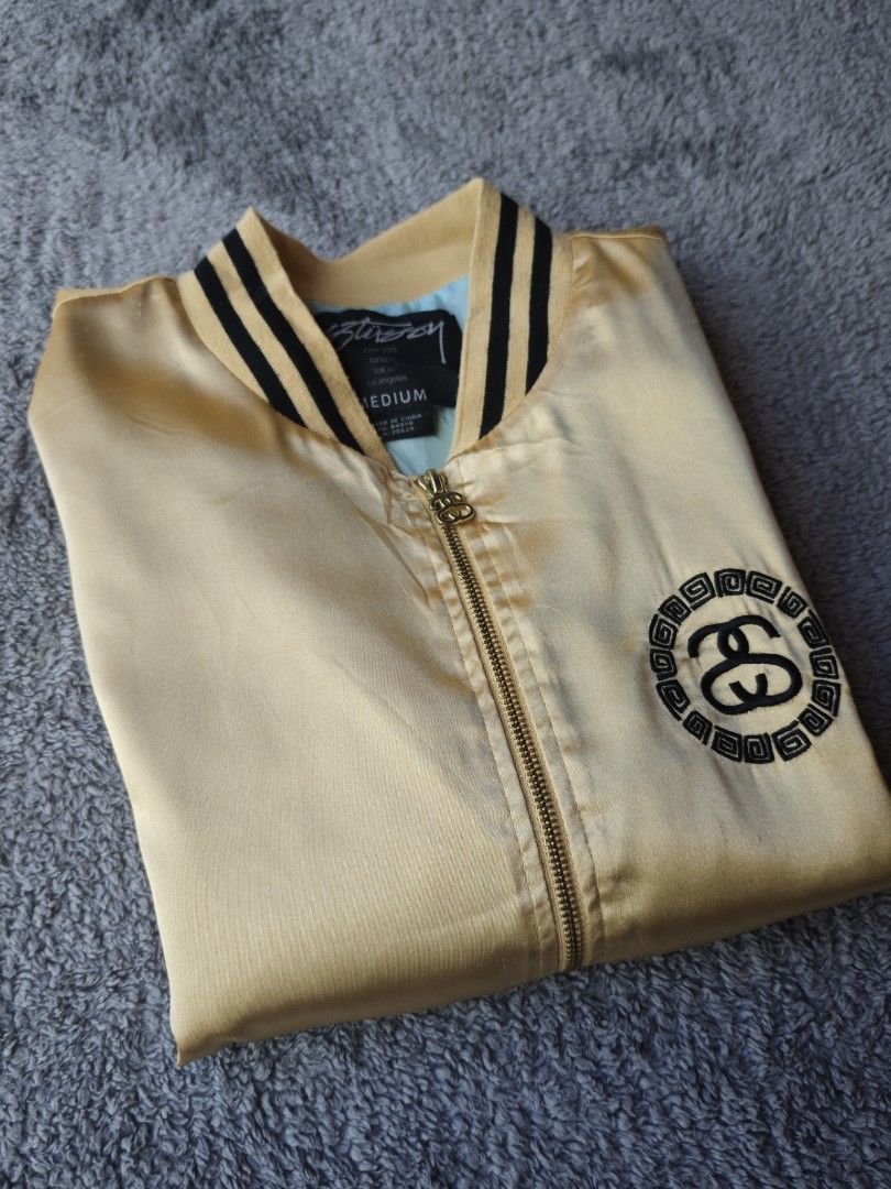 Stussy Satin Bomber Jacket Gold Size Medium (Category: Coats, Jackets and Outerwear) located in Dalaguete. Submitted by three7archives (Listing ID 1469071703, Image 1). Description: - Brand: Stussy - Item: Gold satin bomber jacket - Size: Medium 21/25 - Details: Black and white striped ribbed collar, cuffs, and hem - Branding: Iconic embroidered double S logo on chest and large version on back - Interior: Bright blue inner lining with branded nec