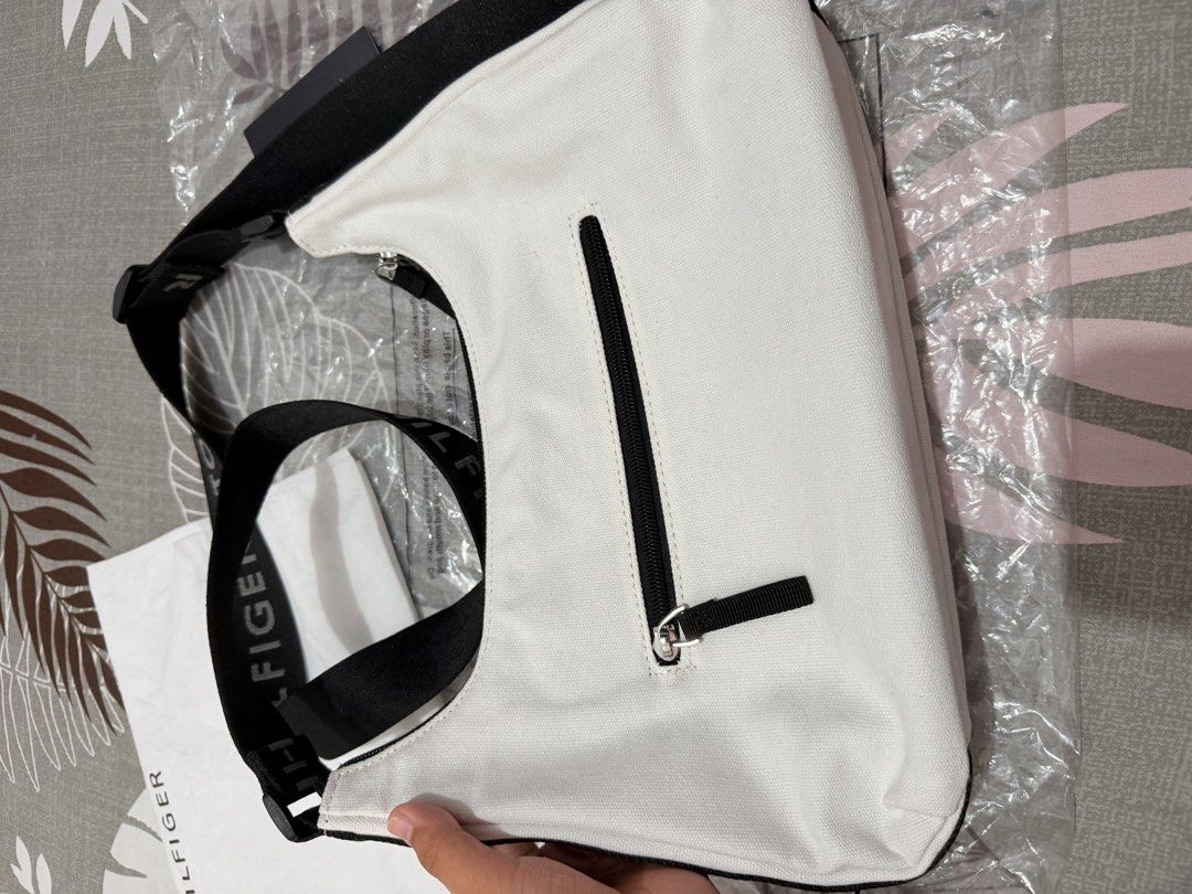 Tommy Hilfiger Sling/Shoulder Bag (Category: Cross-body Bags) located in Munoz. Submitted by shop.holiic (Listing ID 1469099732, Image 3). Description: - Brand: Tommy Hilfiger - Condition: Brand new with tags attached, includes original dust bag - Color: White canvas with black accents - Strap: Adjustable black strap with branding - Pockets: Front patch logo, back zip pocket, inner slip pocket, and inner zip pocket - Interior: Classic blue interi