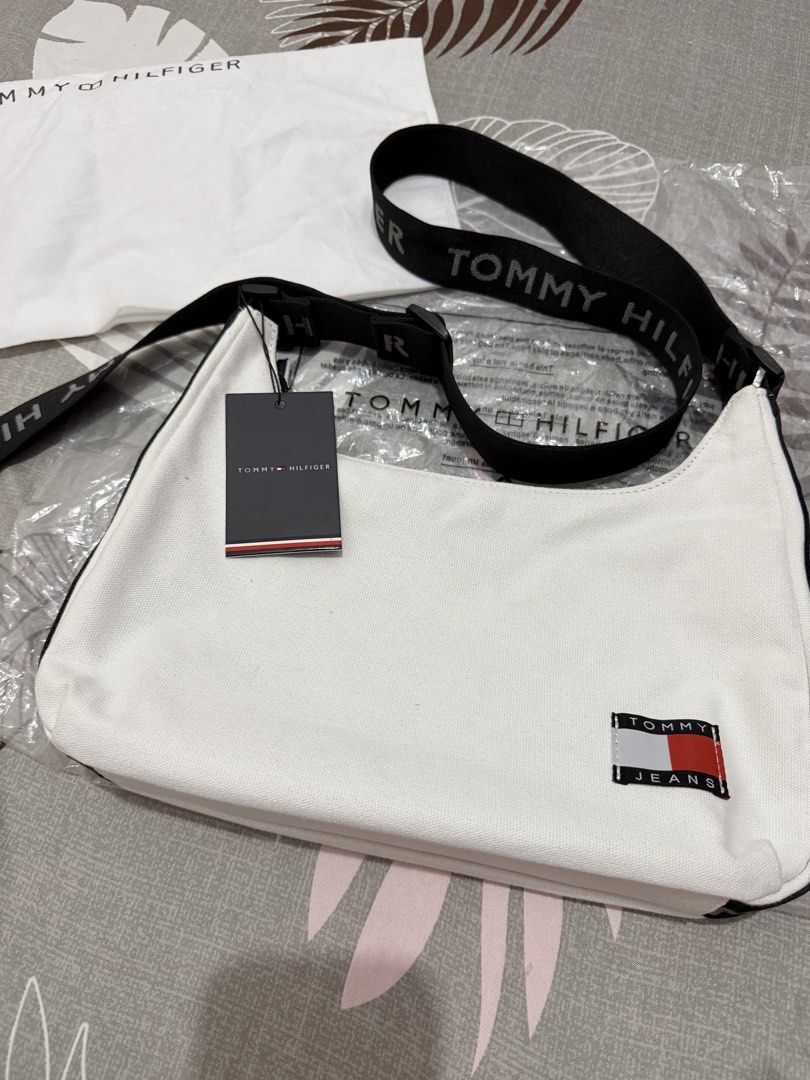 Tommy Hilfiger Sling/Shoulder Bag (Category: Cross-body Bags) located in Munoz. Submitted by shop.holiic (Listing ID 1469099732, Image 1). Description: - Brand: Tommy Hilfiger - Condition: Brand new with tags attached, includes original dust bag - Color: White canvas with black accents - Strap: Adjustable black strap with branding - Pockets: Front patch logo, back zip pocket, inner slip pocket, and inner zip pocket - Interior: Classic blue interi