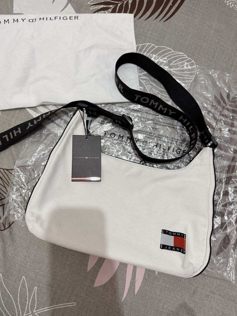 Tommy Hilfiger Sling/Shoulder Bag (Category: Cross-body Bags) located in Munoz. Submitted by shop.holiic (Listing ID 1469099732, Image 2). Description: - Brand: Tommy Hilfiger - Condition: Brand new with tags attached, includes original dust bag - Color: White canvas with black accents - Strap: Adjustable black strap with branding - Pockets: Front patch logo, back zip pocket, inner slip pocket, and inner zip pocket - Interior: Classic blue interi