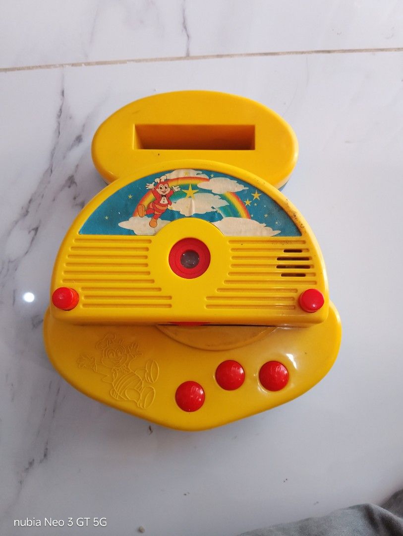 Vintage Jollibee Toy Projector with Slides (Category: Kids Toys) located in Manila. Submitted by wilmen14 (Listing ID 1468949193, Image 3). Description: - Vintage Jollibee toy projector in yellow and blue - Features a classic mascot graphic with a rainbow and stars - Includes two slide cartridges (blue and red) - Requires 3 batteries for operation - Shows minor signs of wear and age on stickers and plastic - Perfect collectible piece for Jollibee