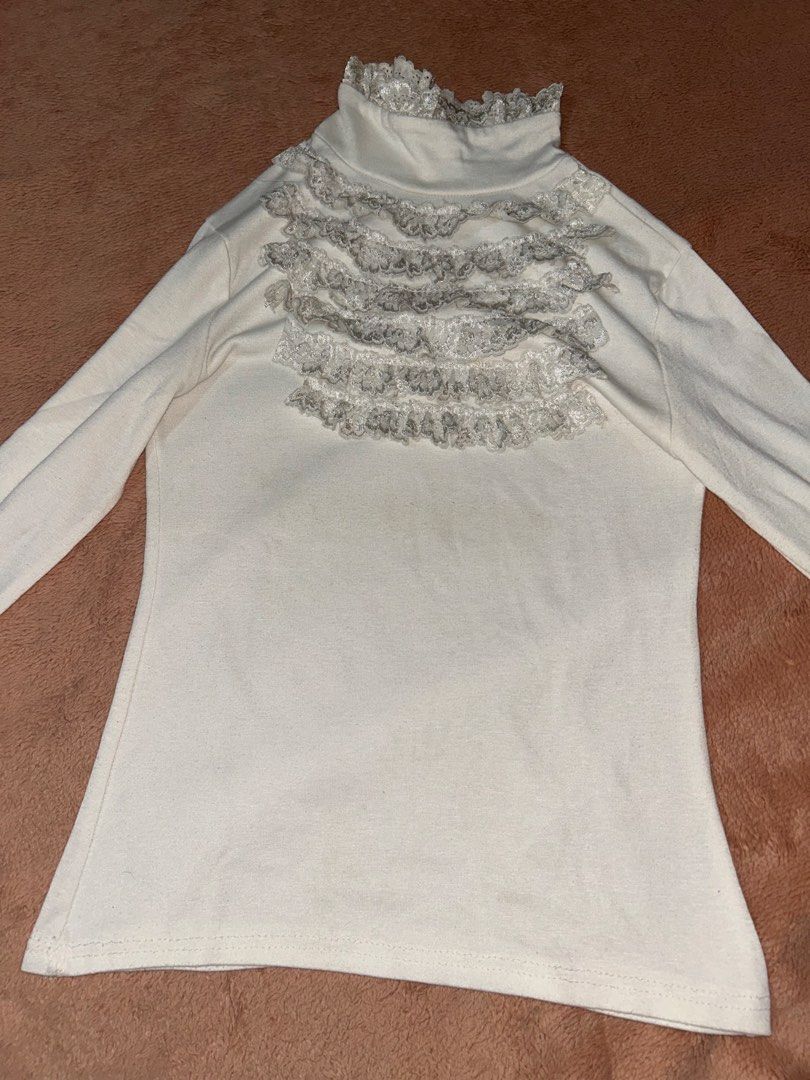 White Turtleneck Long Sleeve Ruffle Lace Top (Category: Longsleeves) located in Manila. Submitted by prelovedbyanavi (Listing ID 1468959078, Image 1). Description: - Color: White with grey lace ruffles across the chest and collar - Fit: Fitted, soft fabric - Style: Long sleeve turtleneck, great for layering under cardigans or jackets .