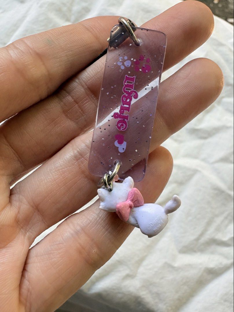 Disney Marie Cat Strap Keychain Charm (Category: Character Goods) located in Tuguegarao City. Submitted by jasminecoll (Listing ID 1465536148, Image 3). Description: - Official Disney Marie cat charm - Features a semi-translucent purple resin tag with glitter, paw prints, and Marie's name - Attached miniature Marie figure with a cute pink bow - Equipped with a black string loop and metal clasp for easy attachment to phones, bags, or keys - Authen