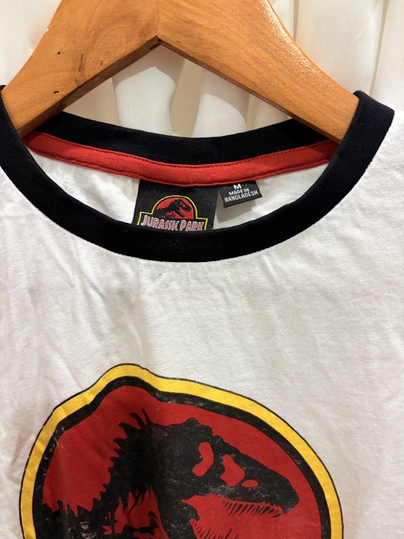 Jurassic Park T-Shirt White Ringer Medium (Category: Tshirts & Polo Shirts ) located in General Trias. Submitted by hayami.clo (Listing ID 1465387103, Image 2). Description: - Item: Jurassic Park Ringer T-Shirt - Size: Medium (tag inside collar) - Color: White with black crew neckline and red interior trim - Design: Distressed classic dinosaur logo on the chest - Fit: Relaxed fit with short sleeves - Origin: Made in Bangladesh - Condition: Clean 