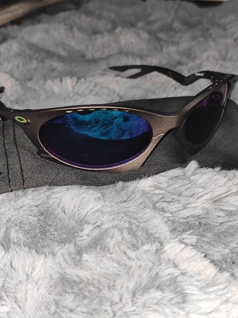 Oakley Sunglasses with Blue Mirrored Lenses (Category: Sunglasses & Eyewear) located in Manila. Submitted by crazylocc887246 (Listing ID 1465487952, Image 1). Description: - Brand: Oakley sunglasses - Frame: Greyish-brown metallic with a futuristic wrap-around design and top brow vent holes - Lenses: Striking blue-purple mirrored finish - Details: Bright green Oakley icon logo on the temple hinge - Inclusions: Comes with a soft grey microfiber po