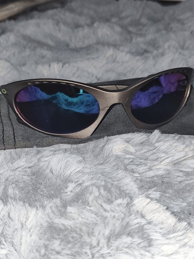 Oakley Sunglasses with Blue Mirrored Lenses (Category: Sunglasses & Eyewear) located in Manila. Submitted by crazylocc887246 (Listing ID 1465487952, Image 2). Description: - Brand: Oakley sunglasses - Frame: Greyish-brown metallic with a futuristic wrap-around design and top brow vent holes - Lenses: Striking blue-purple mirrored finish - Details: Bright green Oakley icon logo on the temple hinge - Inclusions: Comes with a soft grey microfiber po