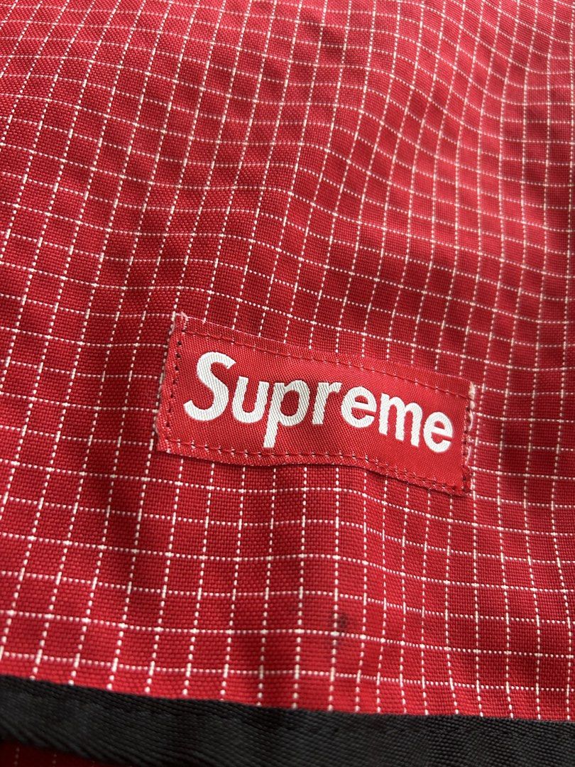 Supreme Red Grid Messenger Bag Made in USA (Category: Sling Bags) located in Cagayan de Oro. Submitted by aljay02 (Listing ID 1465508448, Image 3). Description: Red grid pattern messenger bag from Supreme. White Supreme box logo on the front flap. Adjustable black shoulder strap with silver metal hardware. Dual plastic buckle closure on front straps. Inside tag reads Supreme Made in U.S.A. Measures approximately 18 inches tall. Durable material, 