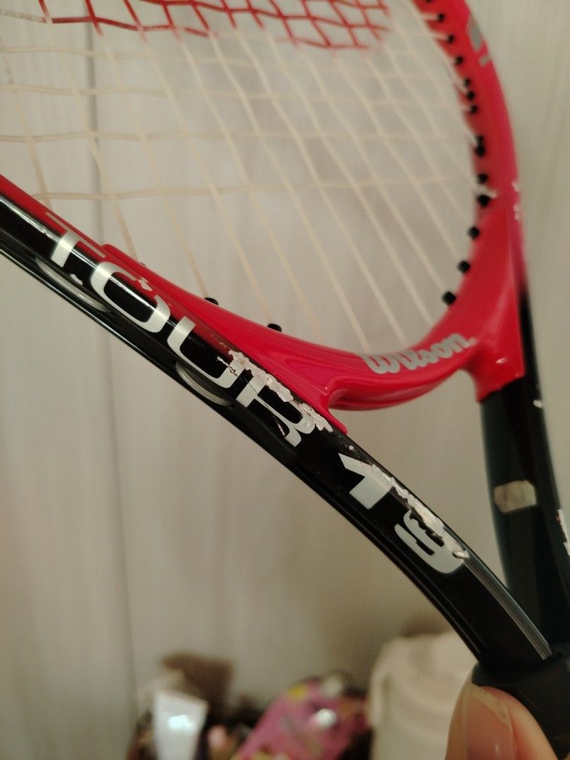 Wilson Tour 19 Tennis Racket (Category: Racket and Ball Sports) located in Tayasan. Submitted by _raceprettyconcept (Listing ID 1465393917, Image 3). Description: - Brand/Model: Wilson Tour 19 tennis racket - Colorway: Striking red and black design - Strings: Strung with white strings featuring the red Wilson logo stencil - Condition: Frame shows noticeable paint chips and scratches from regular court play; grip handle is wrapped in worn tape - I