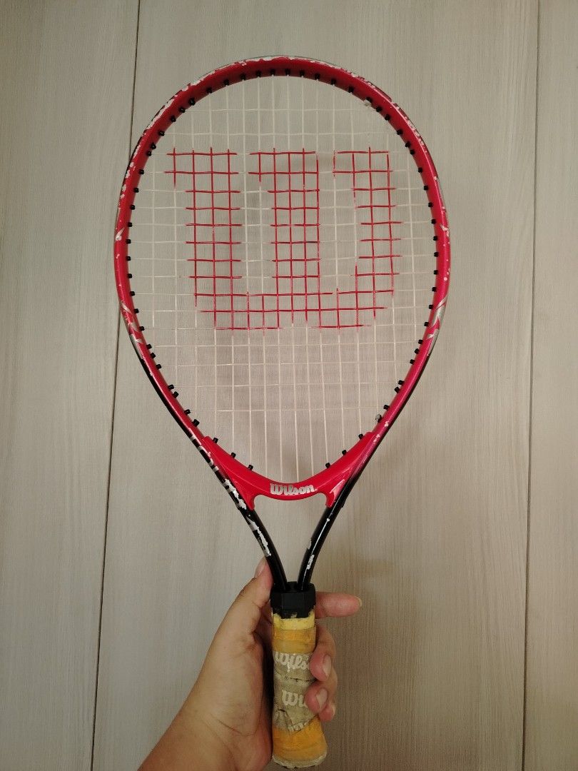 Wilson Tour 19 Tennis Racket (Category: Racket and Ball Sports) located in Tayasan. Submitted by _raceprettyconcept (Listing ID 1465393917, Image 1). Description: - Brand/Model: Wilson Tour 19 tennis racket - Colorway: Striking red and black design - Strings: Strung with white strings featuring the red Wilson logo stencil - Condition: Frame shows noticeable paint chips and scratches from regular court play; grip handle is wrapped in worn tape - I