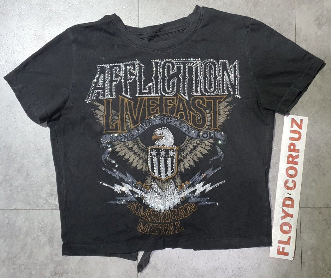 Affliction Live Fast Rhinestone Eagle Graphic T-Shirt (Category: Shirts) located in Laoag. Submitted by floydg_corpuz22 (Listing ID 1469255065, Image 1). Description: Affliction graphic tee with Y2K rocker vibes, featuring rhinestone accents, an eagle shield graphic, and a tie hem. Washed black with distressed details. Affliction for her Xs 17x18 Vgood to excellent need wash 350+sf.