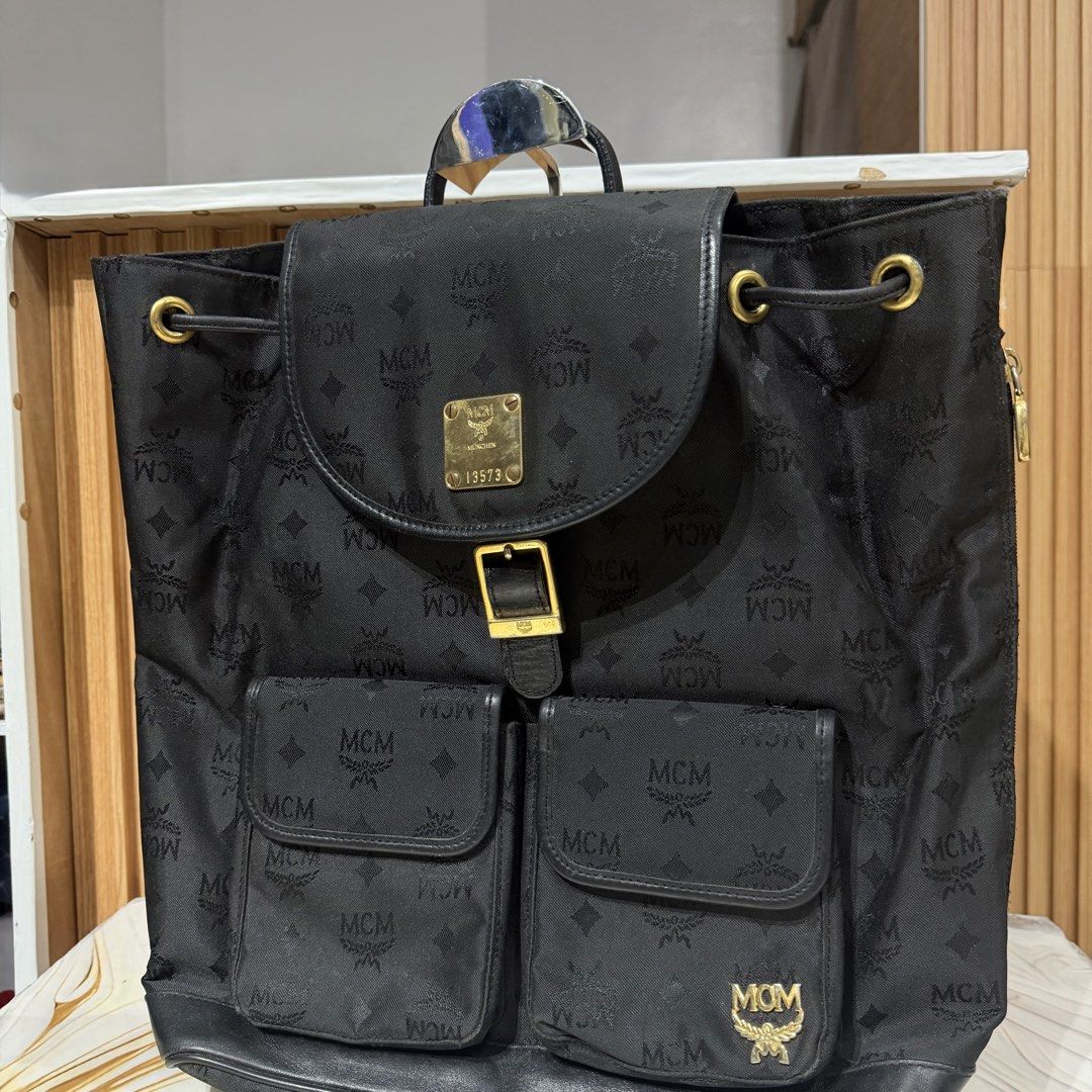 Authentic MCM Black Monogram Nylon Backpack München (Category: Backpacks) located in Bambang. Submitted by heats777 (Listing ID 1469169876, Image 2). Description: - Brand: MCM - Material: Black monogram fabric with leather trim - Hardware: Gold-tone with minor scuffs - Closure: Drawstring under main flap with buckle - Pockets: Two front utility pockets - Details: Classic MCM metal logo plaque and engraved München serial number plate (13573) - Con
