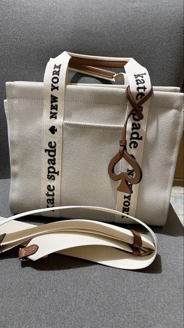 Kate Spade New York Canvas Small Tote (Category: Tote Bags) located in Quezon City. Submitted by bella_ciao (Listing ID 1469260989, Image 1). Description: - Authentic Kate Spade canvas tote bag in neutral cream - Brown leather trim with bold black lettering on the handles - Includes a matching detachable shoulder strap for versatile styling - Features the iconic signature spade charm hanging on the front - Secure zip top closure to keep your belo