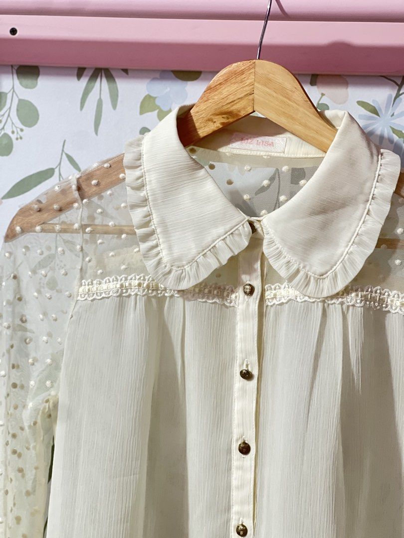 Liz Lisa White Sheer Polka Dot Blouse (Category: Blouses) located in Pasig City. Submitted by morilove (Listing ID 1469162285, Image 2). Description: Liz Lisa cream white blouse with sheer dotted sleeves and yoke. Peter pan collar features delicate ruffles and lace trim across the chest. Button-up front with textured gold-toned buttons. High-low curved hemline. Lightweight chiffon-style fabric on the body. Tag inside the collar..