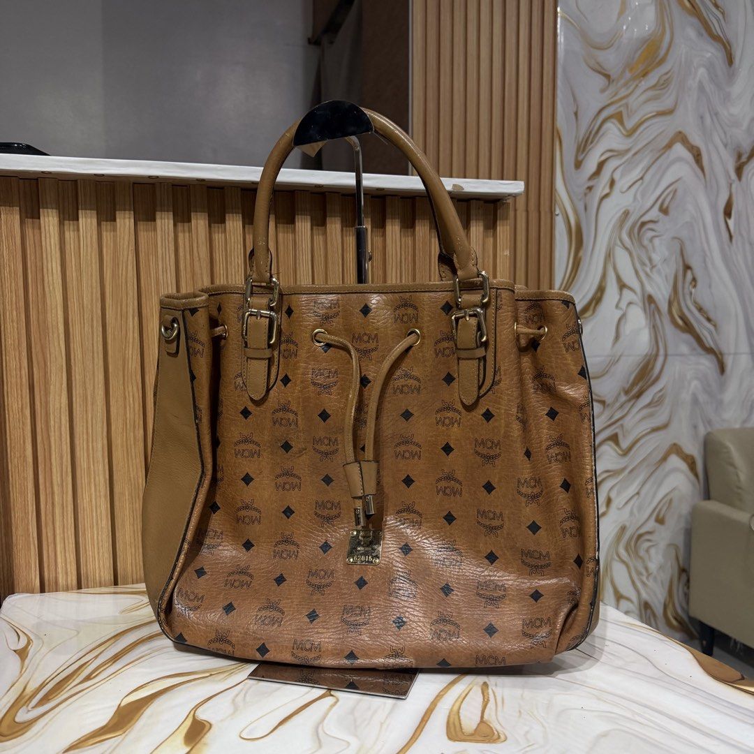 MCM Visetos Brown Monogram Tote Bag (Category: Luxury Bags & Wallets) located in Bambang. Submitted by heats777 (Listing ID 1469169865, Image 1). Description: - Authentic brown MCM Visetos monogram tote bag - Tan leather trim and polished gold-tone hardware - Practical drawstring closure at the front - Double top handles with adjustable buckles - Roomy interior, perfect for daily use - Condition: Regular wear along edges and scuff marks on the bo