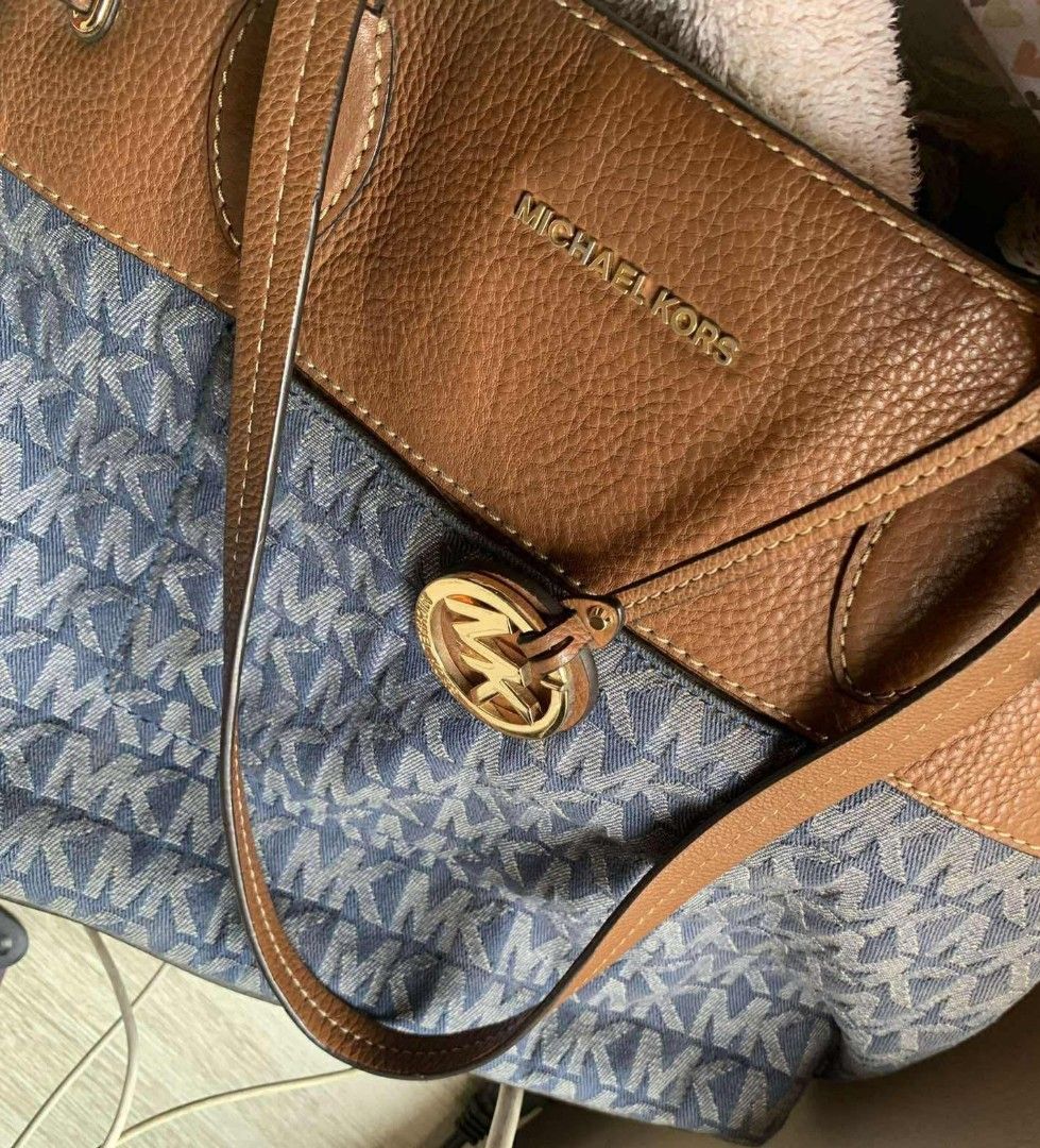 Michael kors mk large denim tote bag (Category: Tote Bags) located in Manila. Submitted by onlinesmartshopping12 (Listing ID 1469380053, Image 1). Description: Stylish Michael Kors tote bag featuring a blue MK monogram pattern with brown leather accents. It has double top handles and a drawstring closure. The interior includes multiple pockets for organization. The bag has a gold-tone MK logo on the front and protective feet on the bottom..