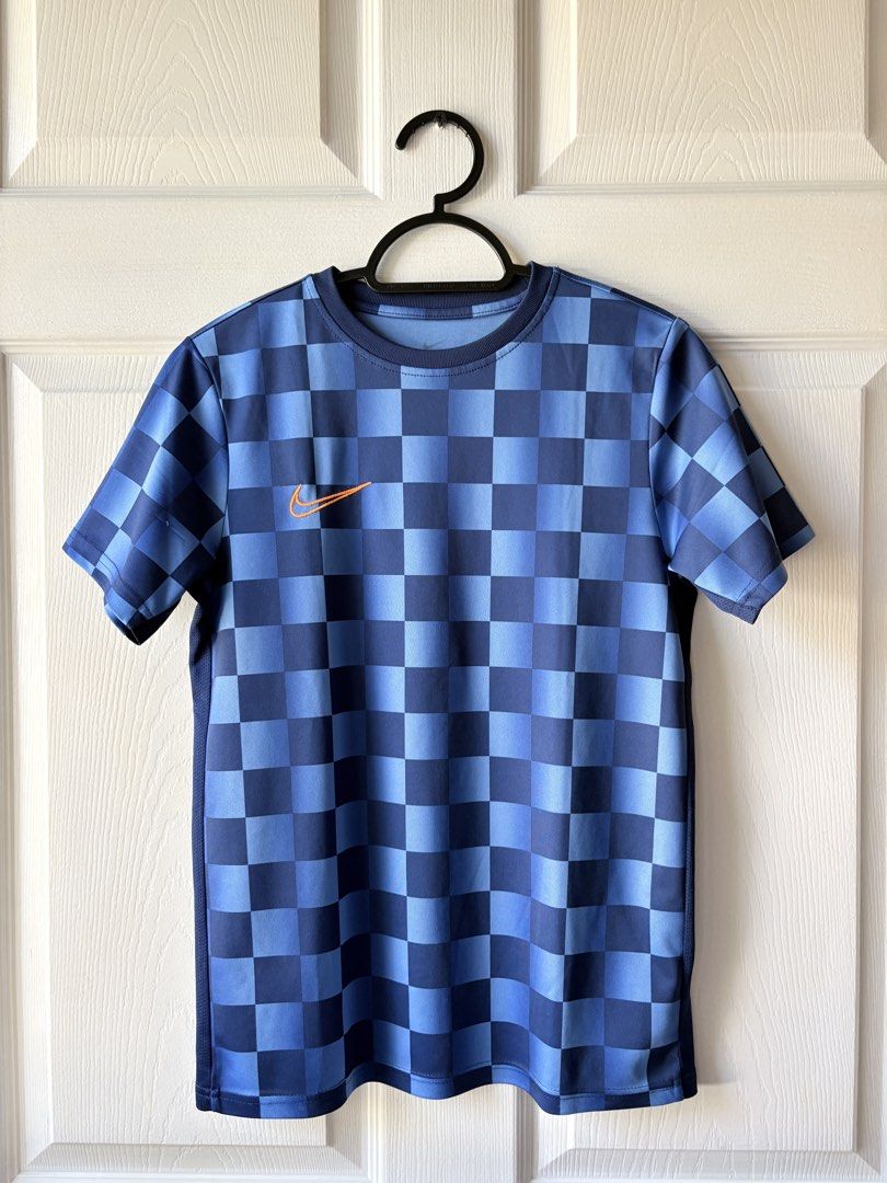 Nike Blue Checkered Football Jersey (Category: Babies & Kids Fashion) located in Liloan. Submitted by mrs.jboiser (Listing ID 1469202940, Image 1). Description: - Brand: Nike - Color: Blue and dark blue checkered pattern - Logo: Orange embroidered Nike swoosh on right chest - Fit: Standard fit jersey with short sleeves and crew neck - Material: Breathable athletic fabric with mesh side panels.