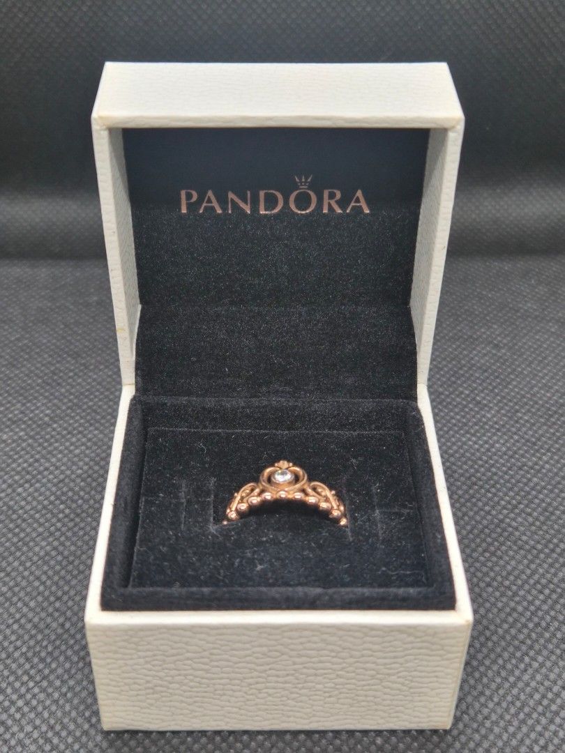 Pandora Rose Gold Princess Crown Ring with Box (Category: Rings) located in Manila. Submitted by prelovedbyc.hannah (Listing ID 1469332191, Image 2). Description: - Brand: Pandora - Material: Rose gold finish - Size: European size 50/ US 5.25-5.5/ 50mm - Design: Princess tiara/crown style with intricate scrollwork and a small clear stone center - Styling: Great for stacking or wearing solo - Inclusions: Original white Pandora box with black velve