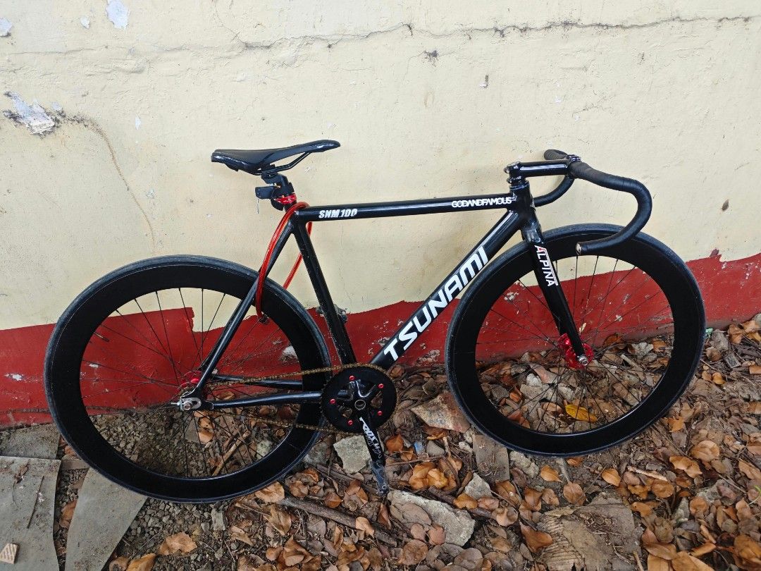 Tsunami SNM100 Fixed Gear Track Bicycle (Category: Bicycles) located in Noveleta. Submitted by johnrenmag03286 (Listing ID 1469175275, Image 1). Description: - Frame: Tsunami SNM100 (Black with white decals) - Handlebars: Drop bars with black tape - Wheelset: Deep section wheels - Drivetrain: Gold chain with red accent bolts on the crankset - Details: Red front hub and red locking cable included - Condition: Solid track setup, ideal for city ridi