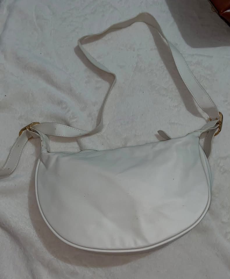 White curved hobo shoulder bag with adjustable strap (Category: Shoulder Bags) located in Manila. Submitted by nobleowl-8fff (Listing ID 1469290706, Image 1). Description: - Color: White - Style: Curved hobo shoulder bag with a clean minimalist design - Material: Smooth finish with structured bottom piping - Hardware: Gold-tone accents on side buckles - Closure: Secure top zipper - Strap: Adjustable long strap for a custom fit - Versatility: Easy