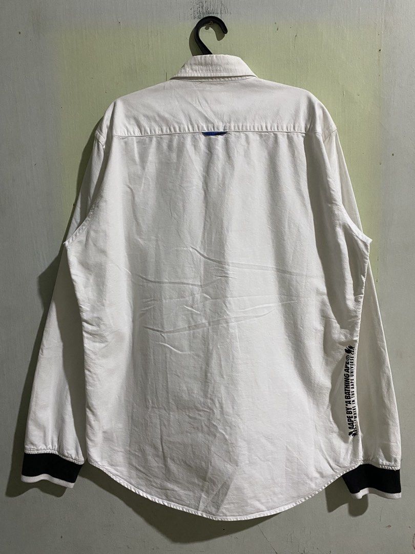 Aape by A Bathing Ape White Button-Down Shirt XL (Category: Tshirts & Polo Shirts ) located in Pandi. Submitted by nasya_thrift_collection2023 (Listing ID 1469545276, Image 2). Description: - Brand: Aape by A Bathing Ape - Color: White with contrast black cuffs - width 22 - length 29 / 30 - Size: XL (fits true to size) - Material: 100% cotton - Details: Black ape logo patch on chest, blue camo lining inside collar, and stylish text prints near he
