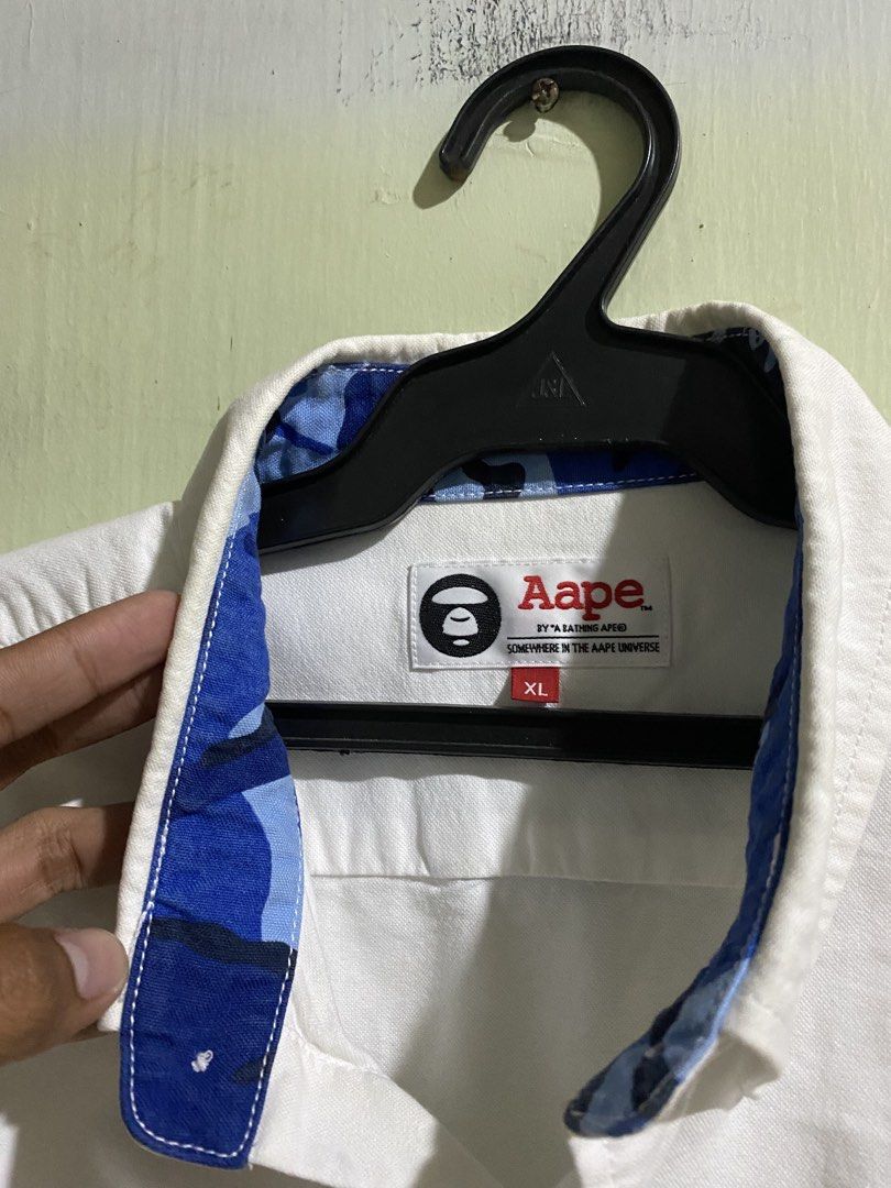 Aape by A Bathing Ape White Button-Down Shirt XL (Category: Tshirts & Polo Shirts ) located in Pandi. Submitted by nasya_thrift_collection2023 (Listing ID 1469545276, Image 3). Description: - Brand: Aape by A Bathing Ape - Color: White with contrast black cuffs - width 22 - length 29 / 30 - Size: XL (fits true to size) - Material: 100% cotton - Details: Black ape logo patch on chest, blue camo lining inside collar, and stylish text prints near he