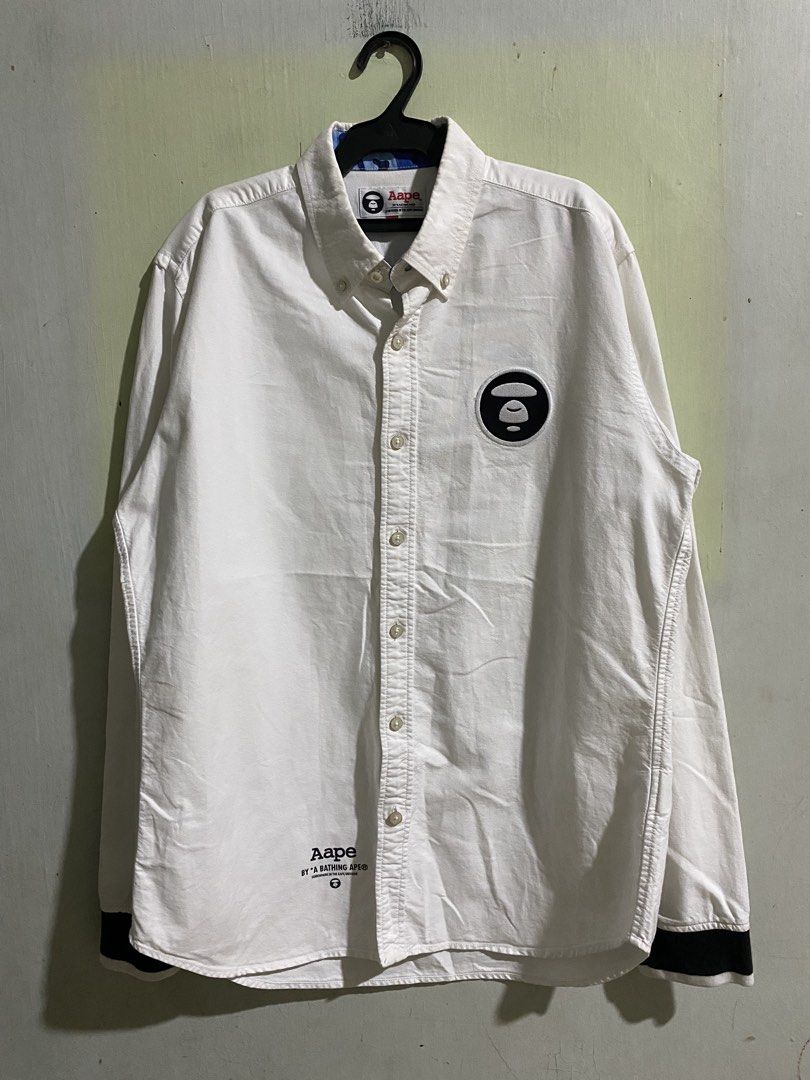 Aape by A Bathing Ape White Button-Down Shirt XL (Category: Tshirts & Polo Shirts ) located in Pandi. Submitted by nasya_thrift_collection2023 (Listing ID 1469545276, Image 1). Description: - Brand: Aape by A Bathing Ape - Color: White with contrast black cuffs - width 22 - length 29 / 30 - Size: XL (fits true to size) - Material: 100% cotton - Details: Black ape logo patch on chest, blue camo lining inside collar, and stylish text prints near he