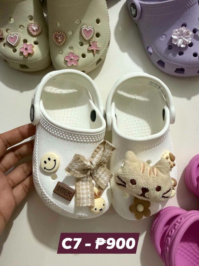 Crocs C7 White Kids Clogs Size C7 with Charms (Category: Babies & Kids Fashion) located in San Fabian. Submitted by hbrst1998 (Listing ID 1469487902, Image 1). Description: - Size: C7 (Kids) - Color: White - Design: Slip-on with secure heel strap - Decorations: Adorned with cute charms (fluffy cat head, checkered bow, smiley face, chocolate bar, and rabbit) - Condition: Clean and well-maintained overall - Perfect fun footwear for daily wear!.
