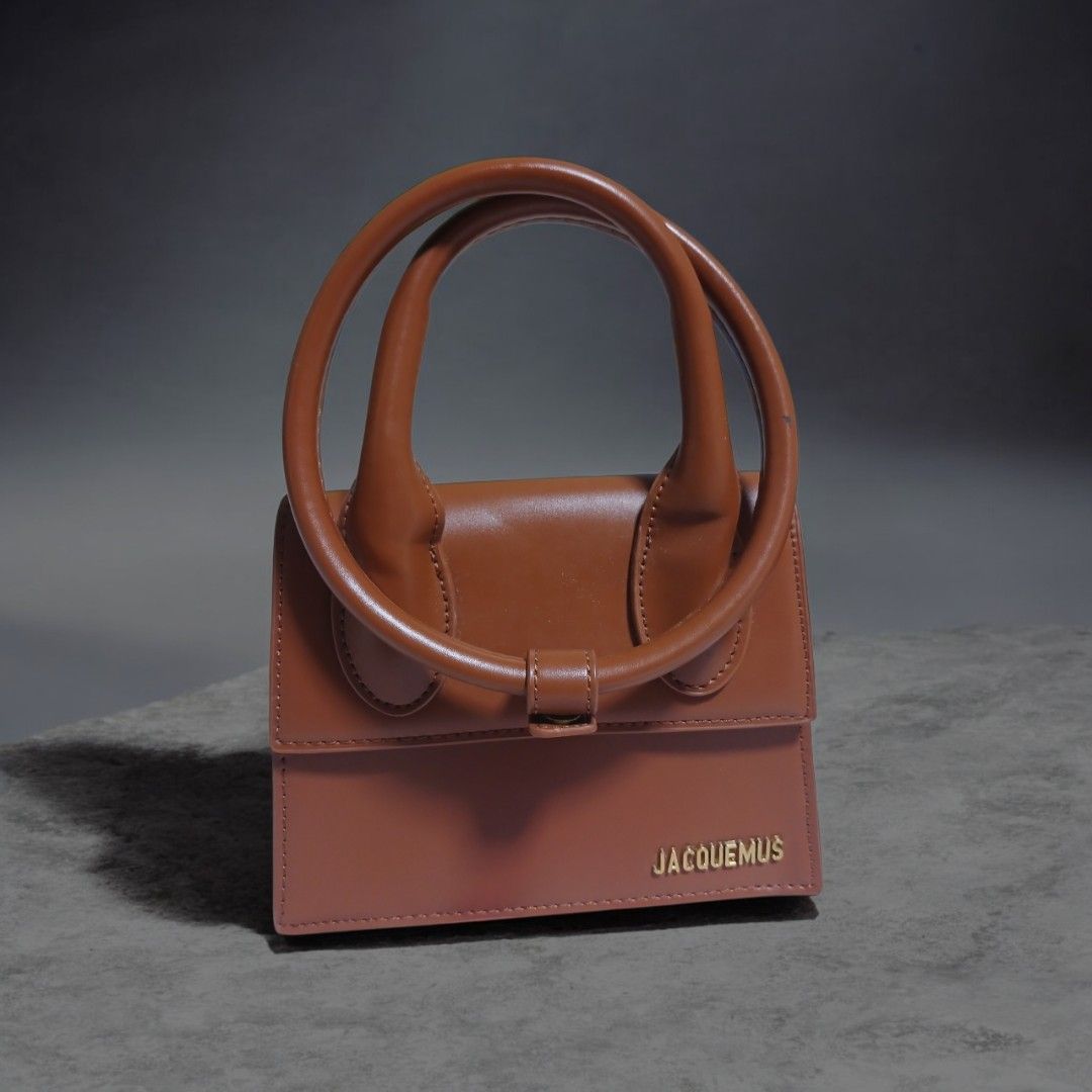 Jacquemus Brown Leather Handbag with Circular Handles (Category: Luxury Bags & Wallets) located in Umingan. Submitted by mrobvariety (Listing ID 1469468680, Image 1). Description: - Brand: Jacquemus - Material: Smooth brown leather with clean edge stitching - Design: Structured boxy shape with gold metallic front lettering - Handles: Double circular rigid top handles - Style: Compact, everyday luxury bag.