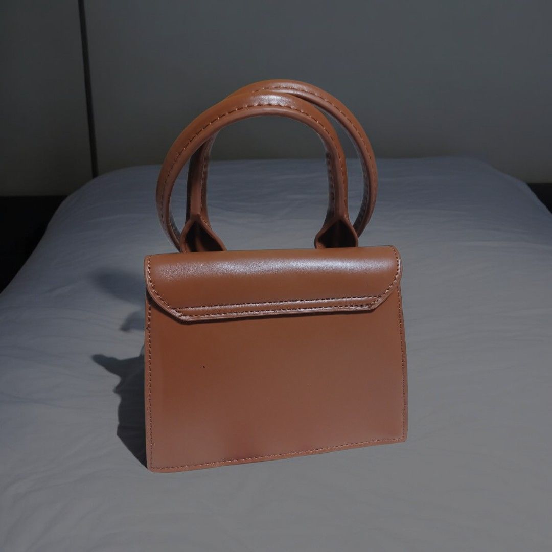 Jacquemus Brown Leather Handbag with Circular Handles (Category: Luxury Bags & Wallets) located in Umingan. Submitted by mrobvariety (Listing ID 1469468680, Image 3). Description: - Brand: Jacquemus - Material: Smooth brown leather with clean edge stitching - Design: Structured boxy shape with gold metallic front lettering - Handles: Double circular rigid top handles - Style: Compact, everyday luxury bag.