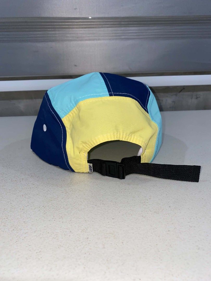 Liefde By Die Dam Duck 5-Panel Cap (Category: Caps & Hats) located in Baler. Submitted by mariokush193815 (Listing ID 1469452559, Image 3). Description: - Brand: Liefde By Die Dam merchandise - Design: Multicolor 5-panel cap with a cute duck graphic on a white front panel - Colors: Contrast navy blue, light blue, yellow panels, and a yellow flat brim - Fit: Adjustable black nylon strap with plastic buckle closure - Details: Interior brand tag and