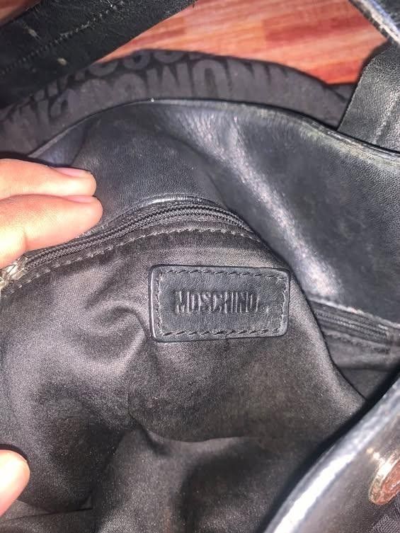 Moschino Black Monogram Shoulder Bag (Category: Luxury Bags & Wallets) located in San Jose del Monte. Submitted by dripfromthrift2 (Listing ID 1469416860, Image 2). Description: - Brand: Moschino - Style: Shoulder bag / Daily tote - Material: Black fabric with all-over monogram logo print and black leather trim - Strap: Adjustable - Hardware: Metal hardware stamped with Moschino branding - Interior: Black fabric lining with an inner zippered pock