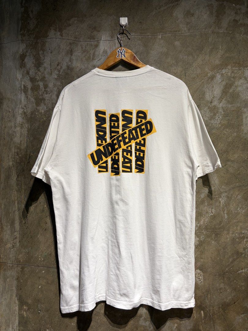 Undefeated White Tee with Yellow Logo Graphic (Category: Tshirts & Polo Shirts ) located in Manila. Submitted by thrifttika (Listing ID 1469535842, Image 1). Description: - Brand: Undefeated - Color: Clean White - Size: 2 - Material: 100% Cotton - Origin: Made in Korea - Design: Large yellow and black back graphic with matching small front chest logo - Condition: Excellent, clean fabric - Style: Simple and versatile streetwear piece.