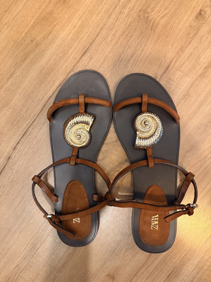 ZARA Brown Seashell Flat Sandals (Category: Flats & Sandals) located in Makati City. Submitted by schelaloo (Listing ID 1469401502, Image 1). Description: - Brand: ZARA - Color: Brown with striking gold seashell hardware - Design: T-strap with adjustable ankle buckle - Soles: Flat black soles with brown heel patch and clear ZARA branding - Versatility: Ideal for daily wear, beach outings, or casual weekend walks.