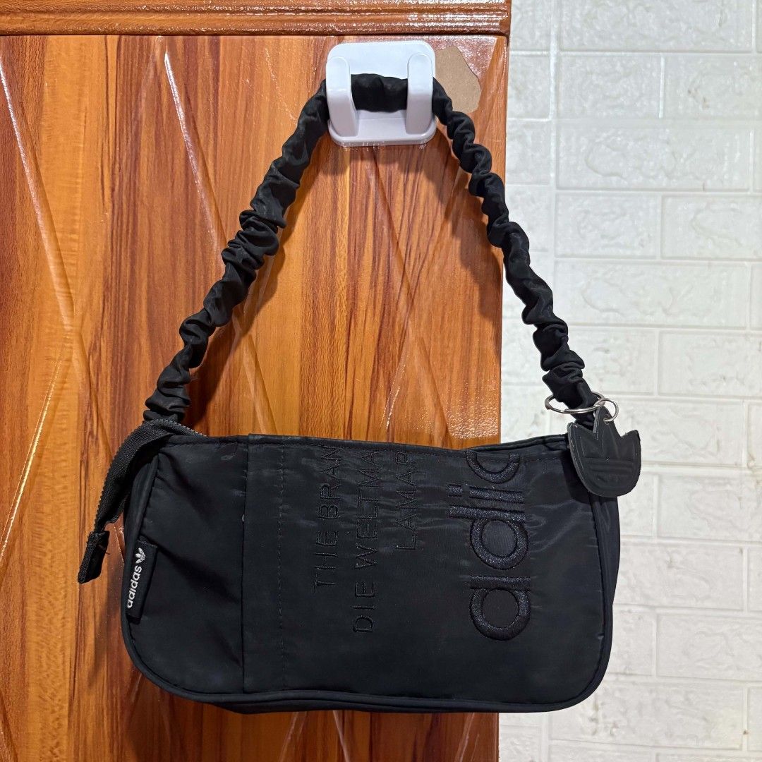 Adidas Black Shoulder Bag Nylon Ruched Strap (Category: Shoulder Bags) located in Carmen. Submitted by ar_thrift00 (Listing ID 1469635000, Image 2). Description: Black nylon shoulder bag from Adidas. Ruched handle strap, zip top closure. Debossed text graphic on front panel alongside Adidas branding tags. Interior lined with zipper pocket and classic Adidas brand patch label. Includes detachable trefoil metal charm pendant on the ring. Compact ba