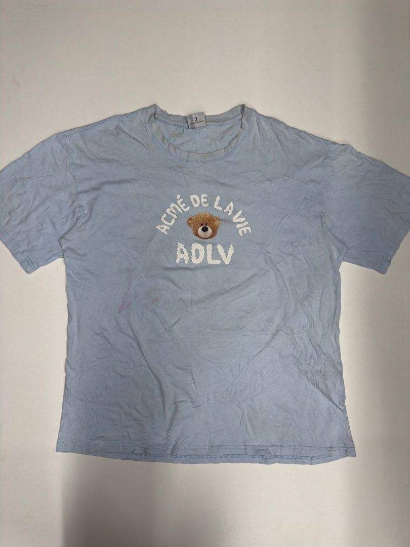 ADLV Light Blue Teddy Bear T-Shirt Size 2 M (Category: Shirts) located in Bustos. Submitted by thenlnclot60500 (Listing ID 1469657274, Image 1). Description: - Brand: Acme de la vie (ADLV) - Design: Teddy bear graphics on front and back - Color: Light blue - Size: Size 2 on tag (equivalent to Medium) - Material: 100% cotton - Origin: Made in Korea - Condition: Normal wear throughout the fabric, with noticeable yellowish stains around the collar, 
