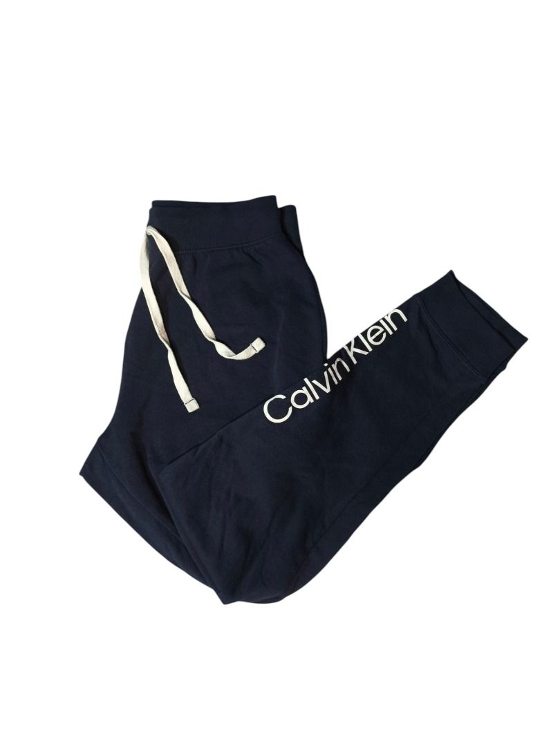 Calvin Klein Navy Logo Sweatpants (Category: Joggers) located in Baguio. Submitted by boneteezandharmony (Listing ID 1469663367, Image 1). Description: Minimalist navy blue Calvin Klein sweatpants featuring bold contrast branding along the leg, cream drawstrings, and ribbed waistband and ankle cuffs. A clean, comfortable piece for casual everyday fits. Tagged Calvin Klein Sleepwear, made in Bangladesh. Condition: 8/10 Dimes: 15 × 39 .