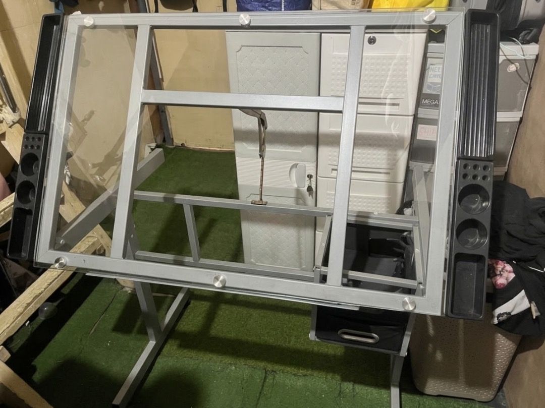 Drafting Glass Table with Extra Side Table Drawers (Category: Tables & Sets) located in Caloocan City. Submitted by elle.23 (Listing ID 1469691732, Image 2). Description: - Adjustable glass top drafting table - Durable grey steel frame with wheels for easy mobility - Includes 2 black fabric storage drawers - Features side storage trays with pen slots and cup holders - Dimensions: approx. 40.9" (W) x 23.6" (D) - Perfect workstation for architects,