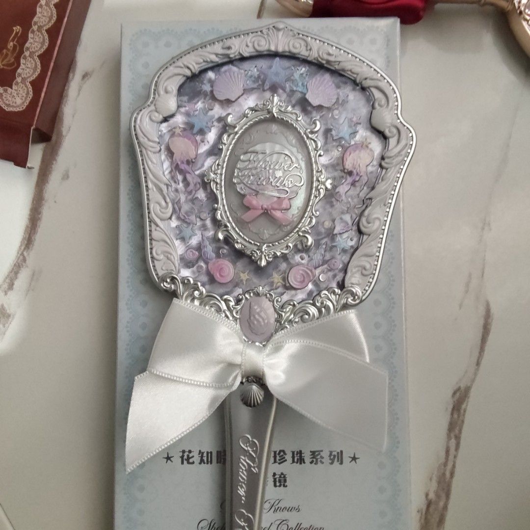 Flower Knows shell jewel handheld mirror - White pearl (Category: Mirrors) located in Manila. Submitted by rosarionav48057 (Listing ID 1469712724, Image 2). Description: - Brand: Flower Knows (shell jewel) - Item: Handheld makeup mirror with real glass surface and intricate central cameo - Design: Ornate silver frame featuring pastel purple and pink details, seashell motifs, and jellyfish designs - Feature: Elegant white satin ribbon tied at the 