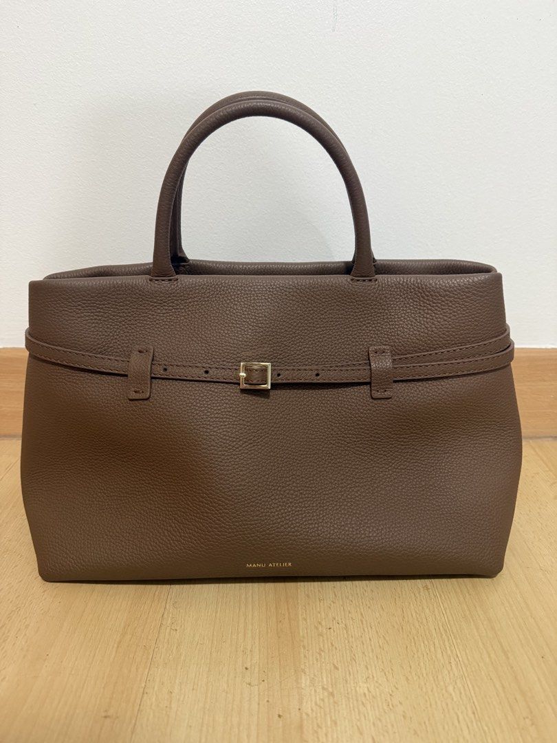 Manu Atelier Brown Leather Tote Bag (Category: Tote Bags) located in Makati City. Submitted by dgcurated09 (Listing ID 1469735988, Image 1). Description: - Brand: Manu Atelier - Material: Premium brown pebbled leather - Design: Structured tote with twin top handles and gold-stamped logo - Details: Decorative buckled strap wrapping around front and back, with protective metal feet underneath - Perfect for daily use, work, or casual outings .