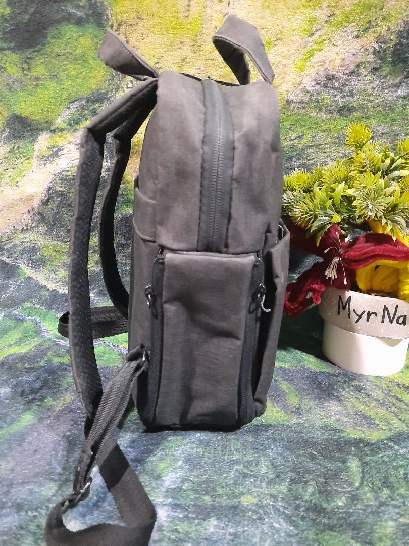 Small camera backpack (Category: Backpacks) located in Baguio. Submitted by klayblakeb87803 (Listing ID 1469678005, Image 3). Description: - Color: Grey - Material: Durable canvas with textured bottom panel - Dimensions: 11" height x 9" base - Compartments: Main zipper compartment and front zippered pocket - Carry options: Dual shoulder straps and top carry handles - Details: Small orange rectangular brand patch on the front - Condition: Clean in