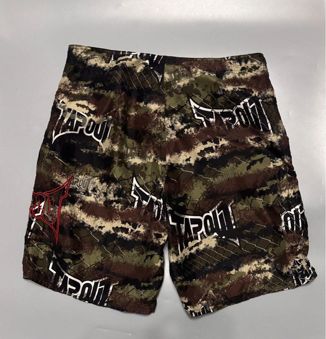 Tapout Camo MMA Board Shorts (Category: Shorts) located in Alaminos. Submitted by outfitcheck.ph (Listing ID 1469698654, Image 2). Description: - Brand: Tapout - Design: Brown, green, and black camo with chain link fence overlay and large white logo - Details: Bright red embroidered logo near the hem - Features: Lightweight fabric, adjustable drawstring waist, and secure side pocket - Condition/Use: Perfect for training, gym sessions, or casual w
