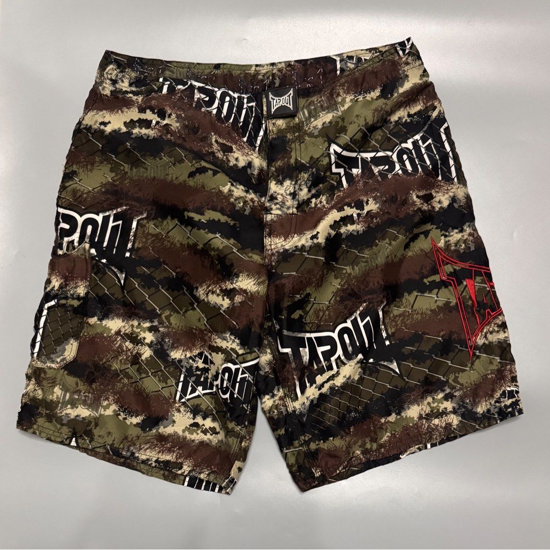 Tapout Camo MMA Board Shorts (Category: Shorts) located in Alaminos. Submitted by outfitcheck.ph (Listing ID 1469698654, Image 1). Description: - Brand: Tapout - Design: Brown, green, and black camo with chain link fence overlay and large white logo - Details: Bright red embroidered logo near the hem - Features: Lightweight fabric, adjustable drawstring waist, and secure side pocket - Condition/Use: Perfect for training, gym sessions, or casual w
