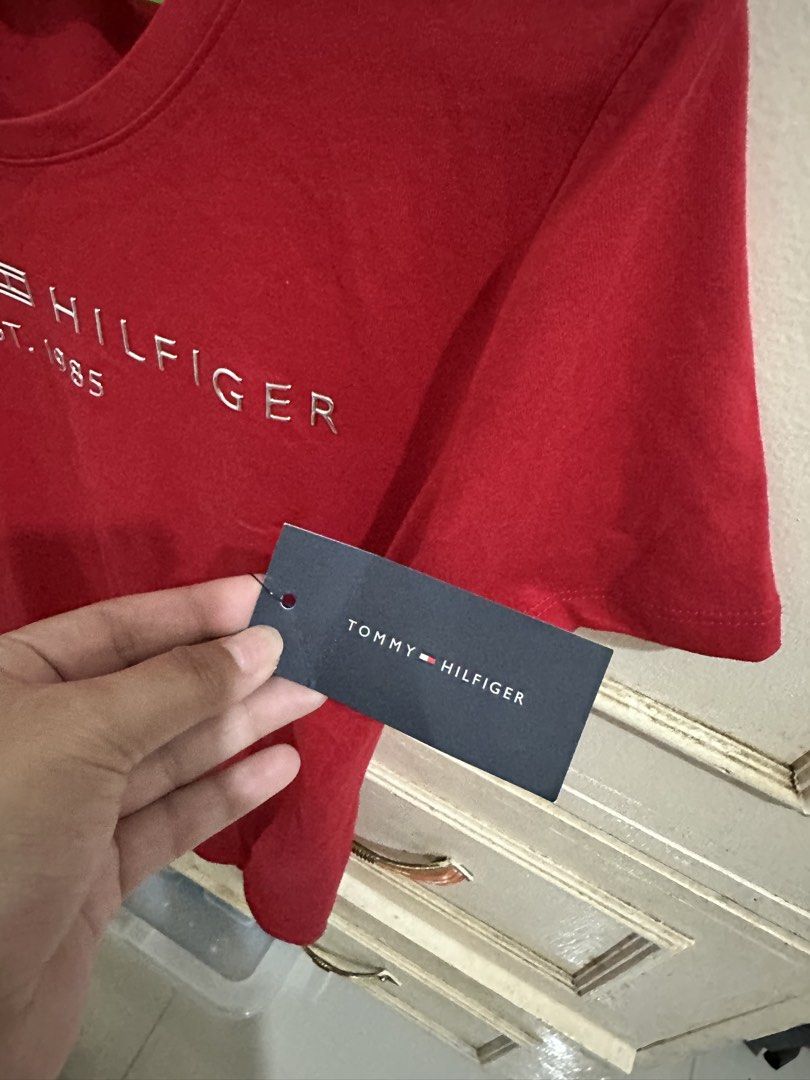 Tommy Hilfiger Red T-Shirt (Category: Shirts) located in Bugallon. Submitted by lavenderlane (Listing ID 1469702945, Image 3). Description: Red short-sleeve t-shirt from Tommy Hilfiger. Bold silver logo embroidery across the chest reads Tommy Hilfiger Est. 1985. Inner collar features striped navy, white, and red detailing. Size PM. Original brand tag attached to the sleeve. Made in Vietnam. Comfortable cotton material..
