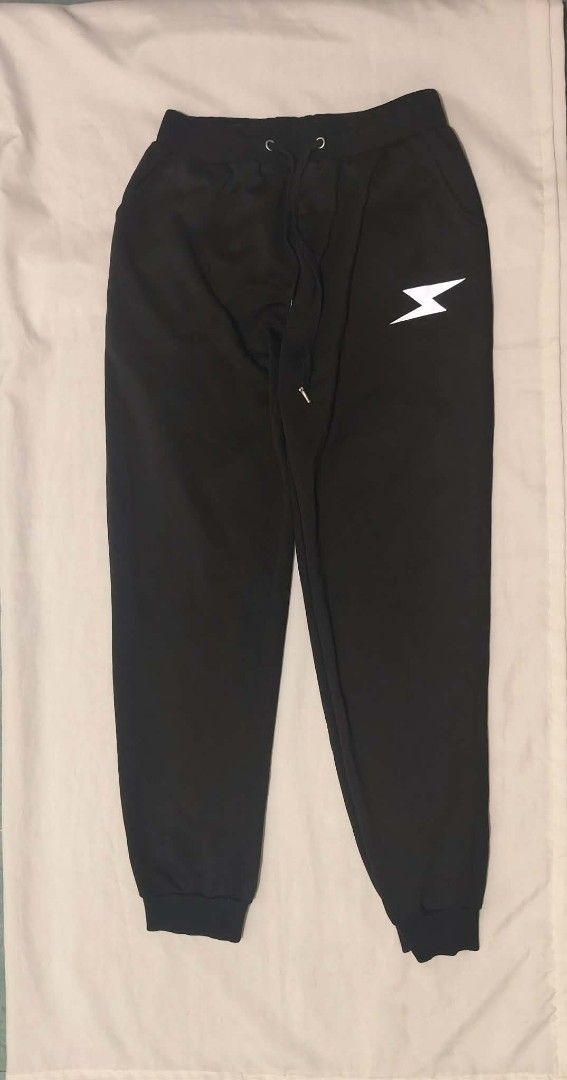 Black Sweatpants With White Lightning Bolt Graphic (Category: Joggers) located in Santa Rosa. Submitted by barbshie (Listing ID 1469826842, Image 1). Description: - Color: Black - Design: White lightning bolt graphic on the left leg - Fit: Relaxed fit with cuffed ankles - Waistband: Elastic with drawstring closures and metal eyelets - Condition: Clean fabric, no stains or damage visible - Best for: Casual wear or lounging at home.