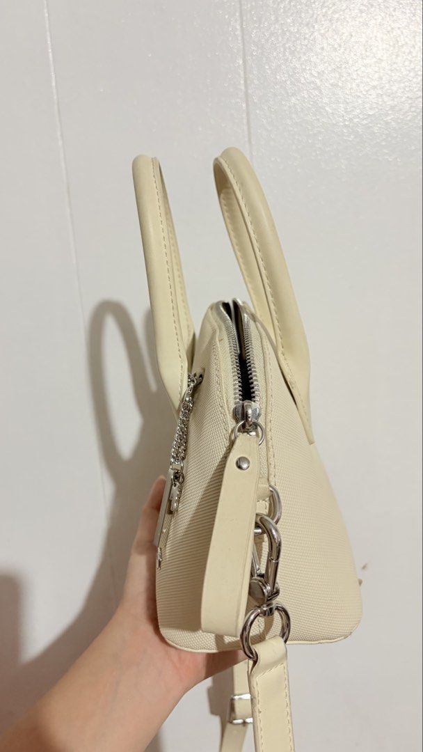 Lacoste Mini Daily Lifestyle Bugatti Strap Bag (Category: Cross-body Bags) located in Baguio. Submitted by meiji50 (Listing ID 1469930651, Image 3). Description: - Brand: Lacoste - Material: Cream textured fabric with black interior lining - Hardware: Silver-tone with zipper closure - Features: Twin top handles, detachable shoulder strap, front crocodile logo plate, branded metal charm, and an interior slip pocket - Condition: Minor wear on the e